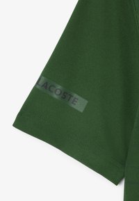 Polo shirt verde a maniche corte con una finitura testurizzata; presenta un pannello trasparente sulla manica con il logo "LACOSTE" in verde scuro.