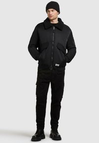 Giubbotto bomber nero con un ampio colletto morbido e chiusura con zip, abbinato a pantaloni cargo neri e stivali neri robusti.