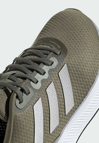 Zapatilla deportiva verde oliva con parte superior texturizada, cordones grises y tres rayas blancas. Cuenta con un logo y una puntera reforzada.