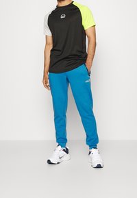 Muž v černém tričku s šedými a neonově zelenými rukávy, modrých teplácích se zipovými kapsami a bílých teniskách Nike stojí na bílém pozadí.