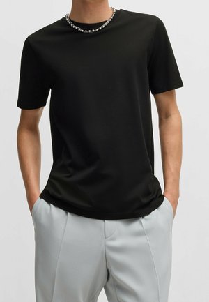 Homme portant un t-shirt noir uni à manches courtes, un pantalon gris clair avec les mains dans les poches, et un collier en perles argentées sur un fond uni.