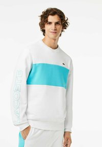 Sudadera blanca con una amplia franja azul aqua en el pecho, con "LACOSTE" en azul claro en la manga. Textura de tela suave.