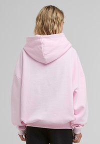 Růžová oversized mikina s kapucí a spadlými rameny, vyrobená z měkkého materiálu a s žebrovanými manžetami. Minimalistický design bez viditelných log.