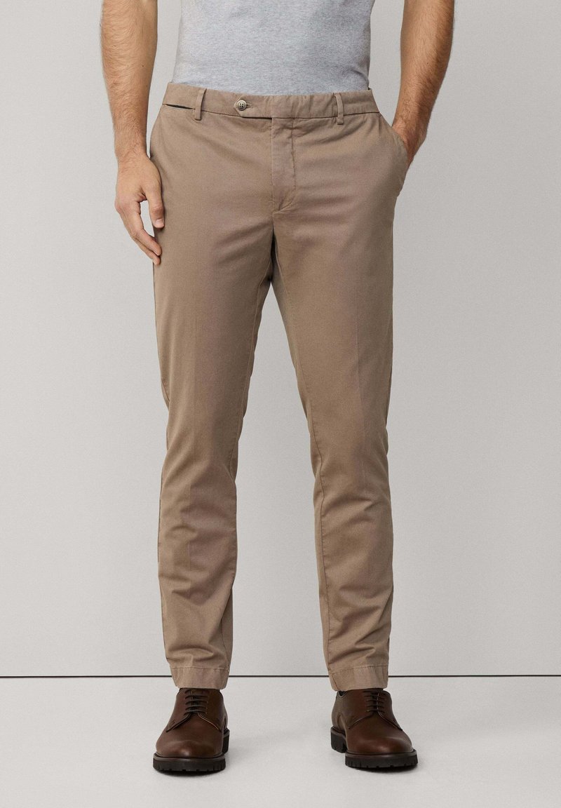 Chinos color khaki realizzati in cotone misto, con frontale piatto, due tasche laterali e chiusura con un solo bottone. Dettaglio della vita regolabile visibile.