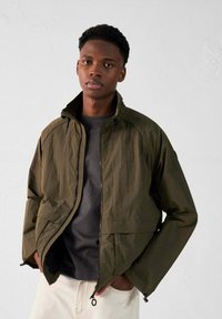 Veste légère olive avec un col haut, fermeture éclair et poches latérales. Portée sur un t-shirt noir avec un tissu texturé.