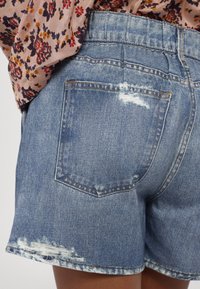 Short en jean avec des détails usés sur la poche arrière et l'ourlet, porté avec un chemisier à motif floral rentré à l'intérieur.