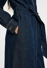 Tummansininen denimtakki vyöllä. Ominaisuuksia ommellut yksityiskohdat, sidottava vyö ja hihansuu metallikoristeella. Pehmeä tekstuuri ja tyylikäs muotoilu.