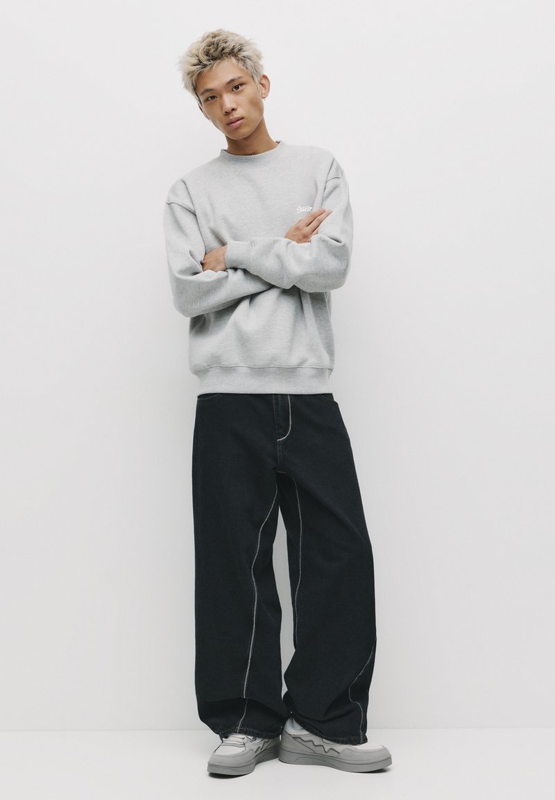 Felpa grigia con polsini a costine, abbinata a jeans a gamba larga blu scuro con cuciture bianche a contrasto. Le scarpe sono grigie chiare con dettagli ondulati.