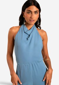 Jumpsuit halter bleu poussiéreux en tissu lisse avec un décolleté drapé. Présente une silhouette ajustée et un design minimaliste.