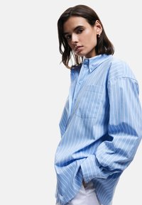 Camicia a righe blu chiaro con bottoni, maniche lunghe, colletto a punta e taschino sul petto, con una vestibilità ampia e rilassata.