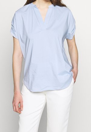 Frau trägt eine hellblaue Kurzarmbluse mit V-Ausschnitt und weiße Hose, eine Hand in der Tasche, steht vor einfachem Hintergrund.