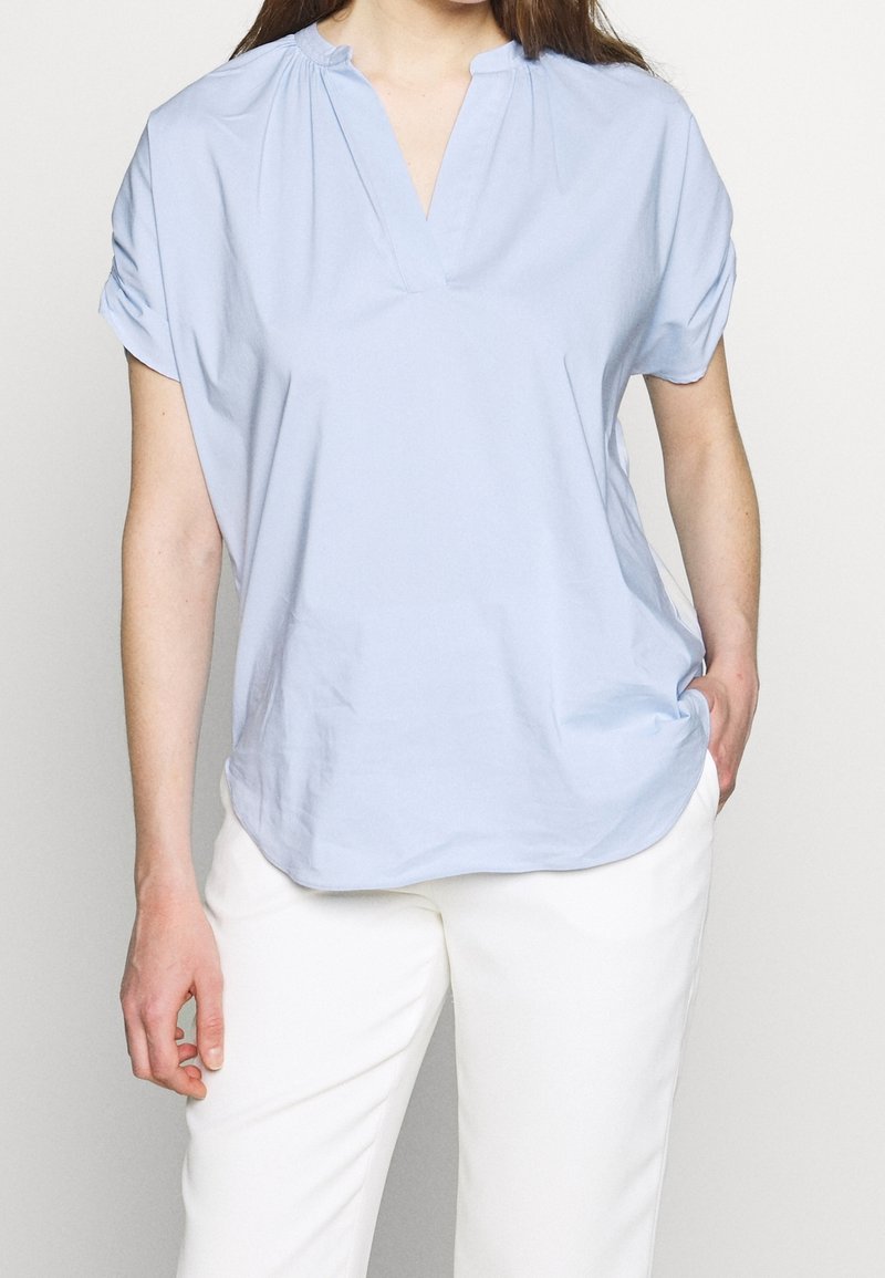Femme portant un chemisier bleu clair à manches courtes avec un col en V et un pantalon blanc, une main dans la poche, debout devant un fond uni.