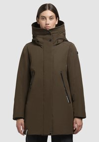 Parka imperméable vert olive avec un col haut, comprenant des poches zippées noires, un devant boutonné et une texture lisse et mate.
