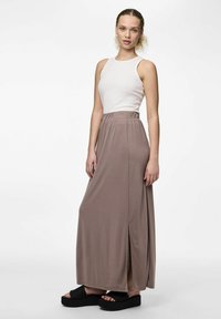 Maxikjol i mjukt taupe-tyg med hög midja och sidoflikdesign, kombinerad med en vit ärmlös topp och klumpiga svarta sandaler.