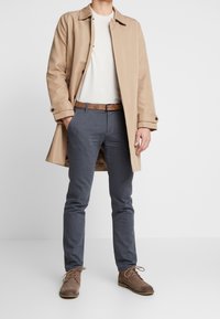 Beige trenčkot přes bílou košili, šedé slim kalhoty a hnědé semišové boty. Mezi pozoruhodné detaily patří texturovaný pásek a knoflíkové manžety kabátu.