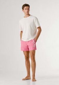 Jonge man die blootsvoets staat, gekleed in een effen wit T-shirt en roze korte broek met witte letters aan de zijkant.