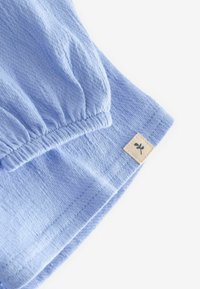 Himmelblaues Baumwollgewebe mit geraffter Textur, versehen mit einem gestickten Saum und einem kleinen gewebten Etikett in Beige mit einem schwarzen Emblem.