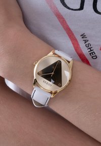 Gouden en zwarte driehoekige wijzerplaat horloge met een witte leren band. Bevat minimalistische uurmarkeringen en een metalen kroon aan de zijkant.