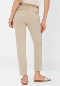 Beige, enganliegende Hose mit dezenter Glanzoptik, ausgestattet mit Gesäßtaschen und schmalem Beinschnitt. Kombiniert mit einem hellen Oberteil und offenen Sandalen.