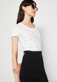 Hvid t-shirt med korte ærmer, der har et diskret mønster på forsiden, parret med en sort ribbet nederdel, der viser CK-logoet på taljebåndet.