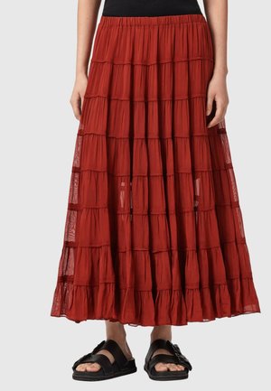 Kobieta ubrana w długą, warstwową, rdzawe-czerwoną spódnicę z przezroczystymi panelami oraz czarne sandały z klamrami, stojąca na tle jednolitego tła.