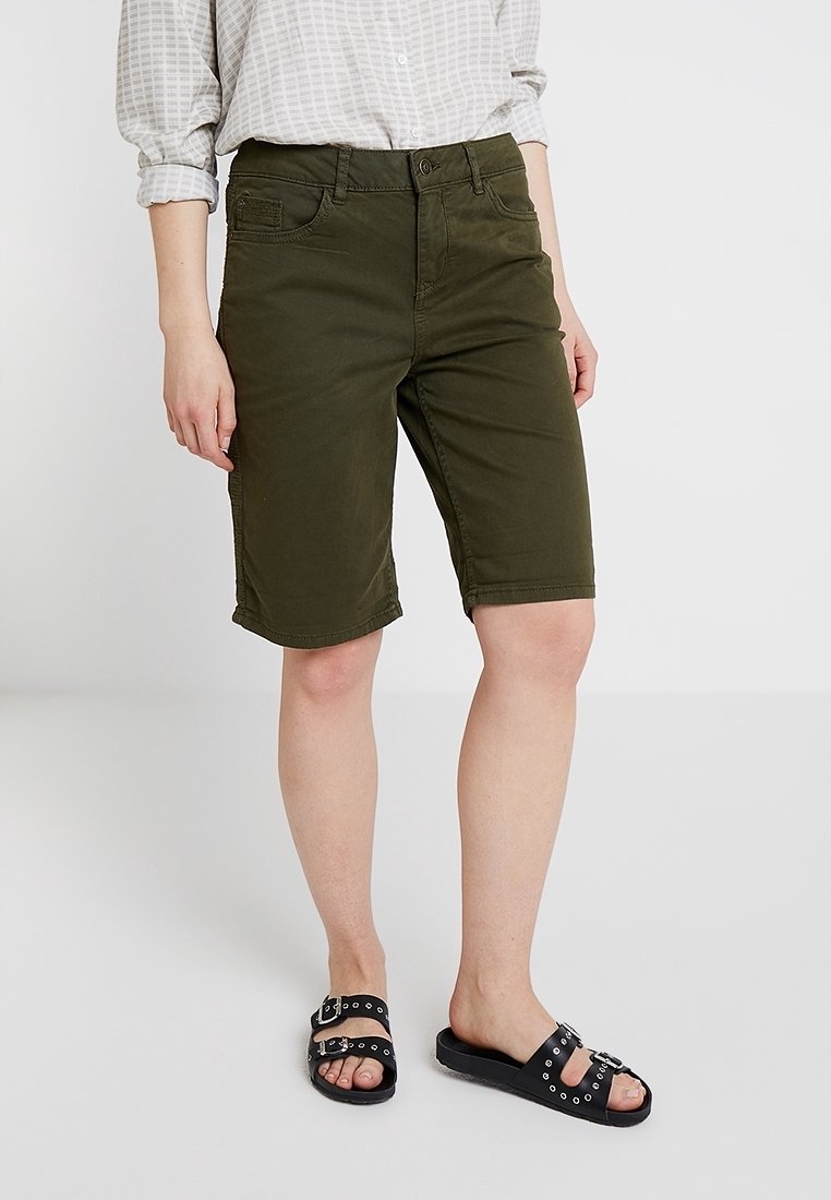 Bermudas en mélange de coton vert olive avec une fermeture à bouton à l'avant, des passants de ceinture, ainsi que des poches latérales et arrière, assorties à des sandales noires à enfiler.
