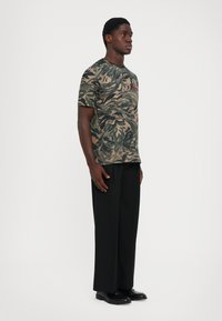 Hombre de pie con pantalones negros y zapatos negros, vistiendo una camiseta de camuflaje verde y marrón con un diseño de cara de tigre.