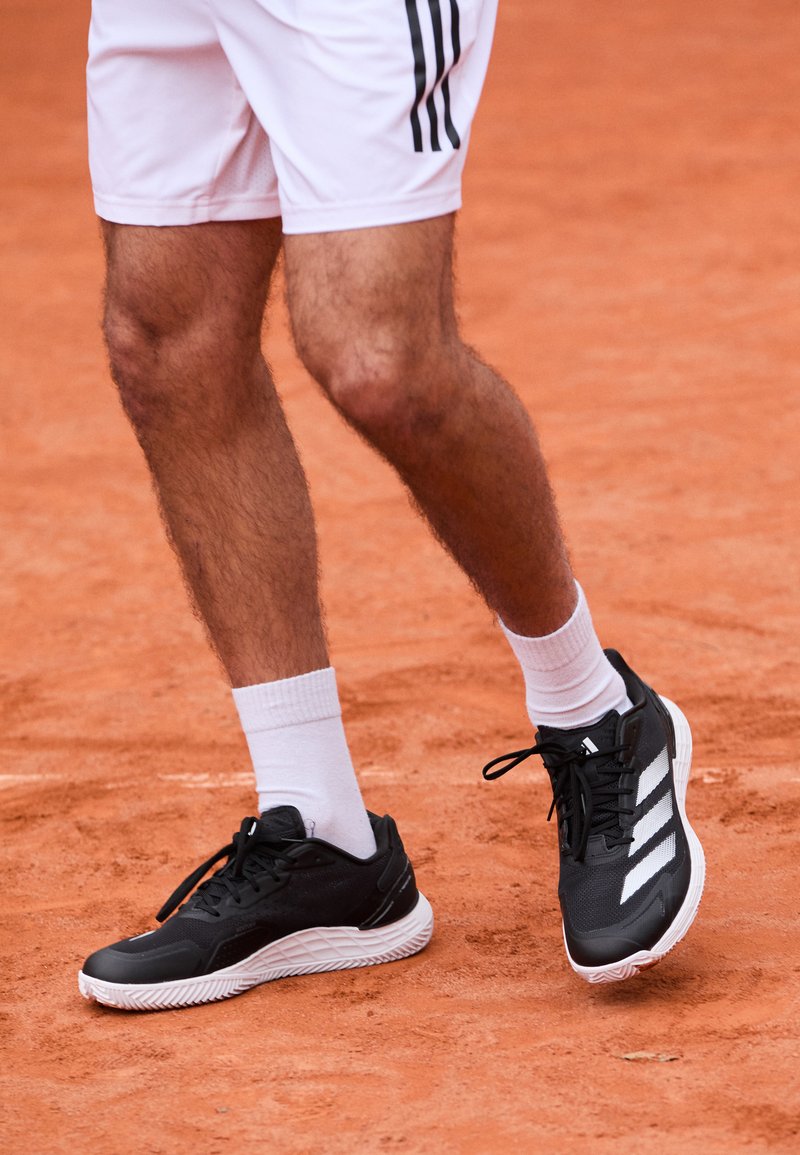 Ben på en person som har på seg hvite shorts, hvite sokker og svarte Adidas-sko, stående på en leir tennisbane.