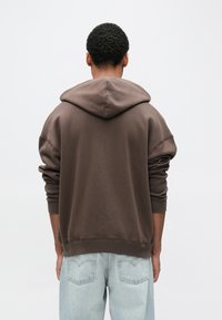 Hnedá nadrozmerná mikina s kapucňou z mäkkej látky, s veľkou kapucňou a zužujúcimi sa rukávmi. Kombinovaná so svetlomodrými džínsami.