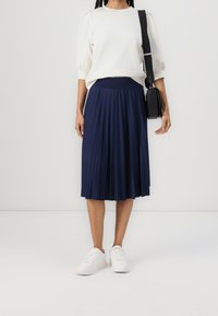 Jupe midi plissée bleu marine avec une taille lisse, associée à un pull crème et des baskets blanches. Un sac bandoulière noir apporte un détail contrastant.