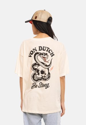 Femme portant un t-shirt beige Von Dutch avec un motif au dos représentant un serpent enroulé autour d’un crâne et le texte « Be Strong », associé à une casquette beige.