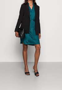 Tyrkisgrøn kjole med bælte, lagdelt under en sort blazer. Har V-hals og flæsekant. kombineret med sorte åbne hæle og en quiltet taske.