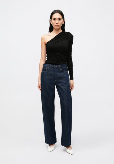 Čierny top na jedno rameno z textúrovanej látky, skombinovaný s tmavomodrými širokými džínsami s vysokým pásom a bielymi lodičkami s špicatou špičkou.