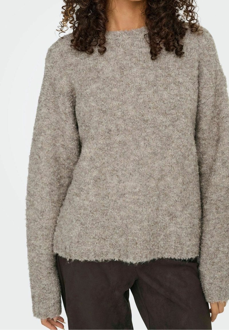 Femme portant un pull à manches longues en maille douce gris et un pantalon foncé, avec des cheveux bouclés partiellement visibles au niveau du cou.