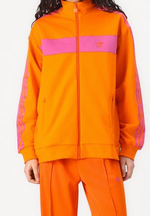 Person trägt einen orangefarbenen Adidas-Trainingsanzug mit pinkfarbenen Streifen an den Ärmeln und einem pinkfarbenen Querband über der Brust.