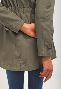 Olivově zelená bunda s úzkým pasem, knoflíkovými manžetami a hladkým povrchem. Nositá přes modré džíny.