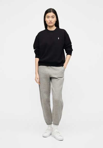 Γυναίκα που στέκεται φορώντας μαύρο πουλόβερ με μακριά μανίκια, γκρι φόρμα, λευκές κάλτσες και λευκά αθλητικά παπούτσια, με το ένα χέρι στην τσέπη.