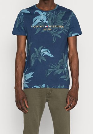 Homme portant un t-shirt Tommy Hilfiger bleu marine avec des imprimés de feuilles tropicales bleu clair et un pantalon vert olive.