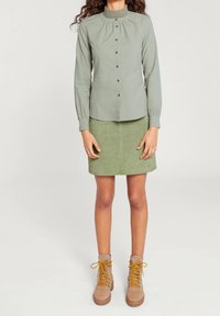 Vaaleanvihreä nappipaitapusero korkealla kauluksella ja pitkillä hihoilla, yhdistetty samanväriseen mokkanahkaiseen minihameeseen ja beigeihin nauhallisiin saappaisiin.