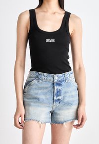 Kvinna iklädd en svart ärmlös topp med vitt logo och ljusblå fransade denimshorts som står mot en enfärgad bakgrund.