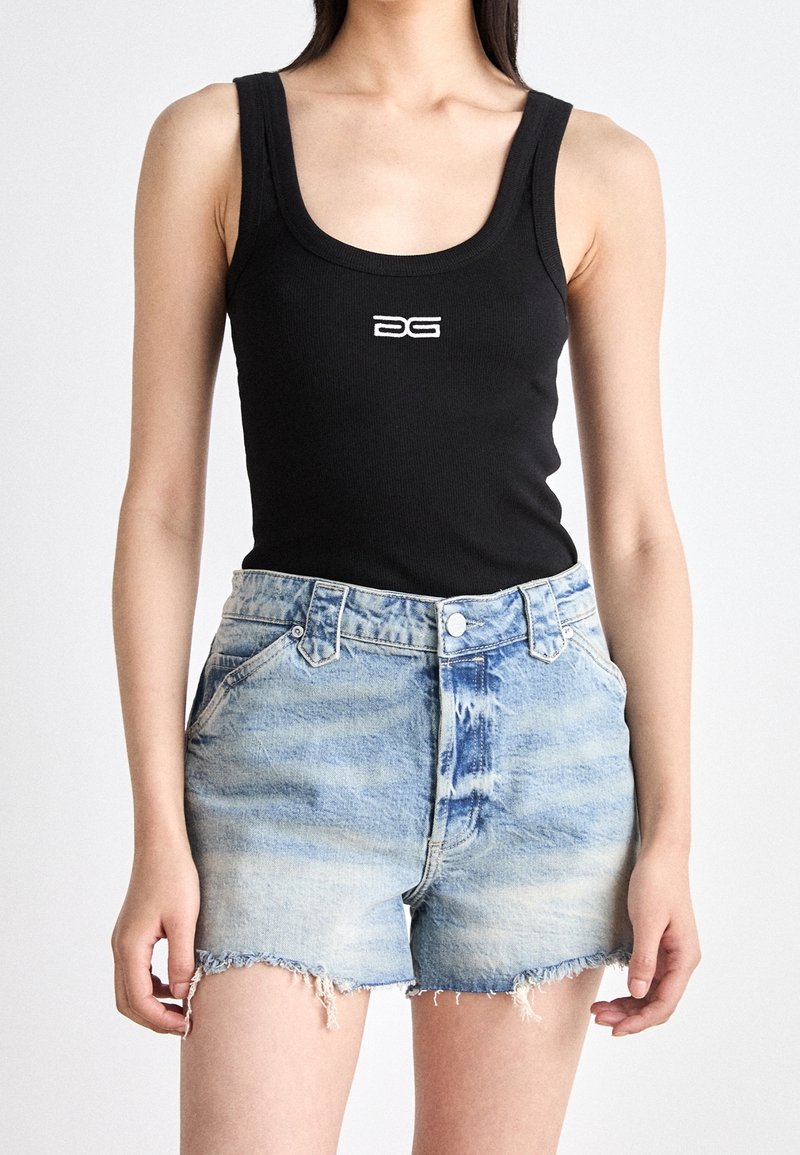 Kvinna iklädd en svart ärmlös topp med vitt logo och ljusblå fransade denimshorts som står mot en enfärgad bakgrund.