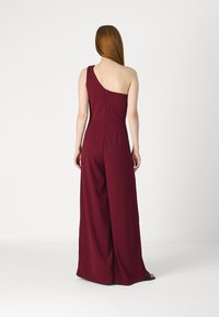 Combinaison rouge bourgogne à une épaule avec un design à jambes larges, texture de tissu lisse et fermeture éclair dans le dos.
