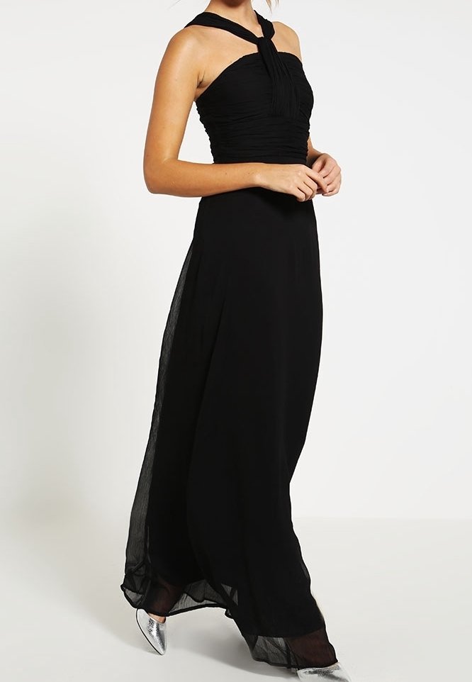 Femme portant une robe de soirée noire sans manches à col halter et des chaussures argentées à bout pointu, debout sur un fond blanc.