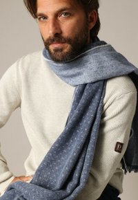 Foulard bleu à motif en tissu doux et texturé drapé sur un pull à côtes de couleur claire, avec une petite étiquette de marque sur la manche.