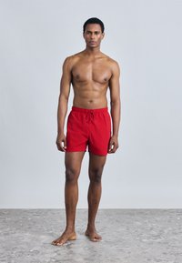 Κόκκινα μαγιό σορτς με ελαστική μέση και κορδόνι, κατασκευασμένα από ελαφρύ ύφασμα. Επίπεδο μπροστινό μέρος και άνετη εφαρμογή, πάνω σε υφασμένο υπόστρωμα.
