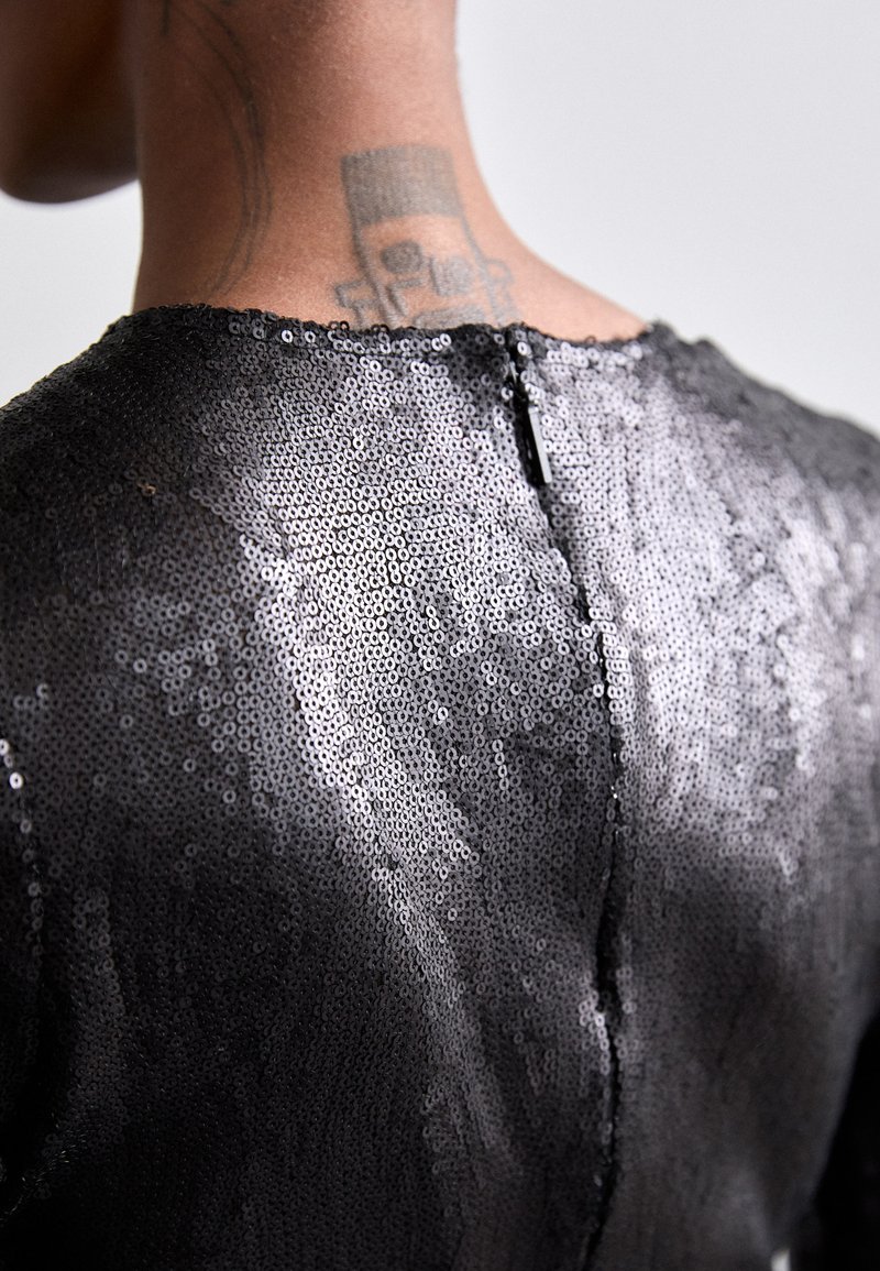 Vêtement en sequins noirs avec une texture lisse et un col haut. Il présente une fermeture zip à l'arrière et un ourlet rond.