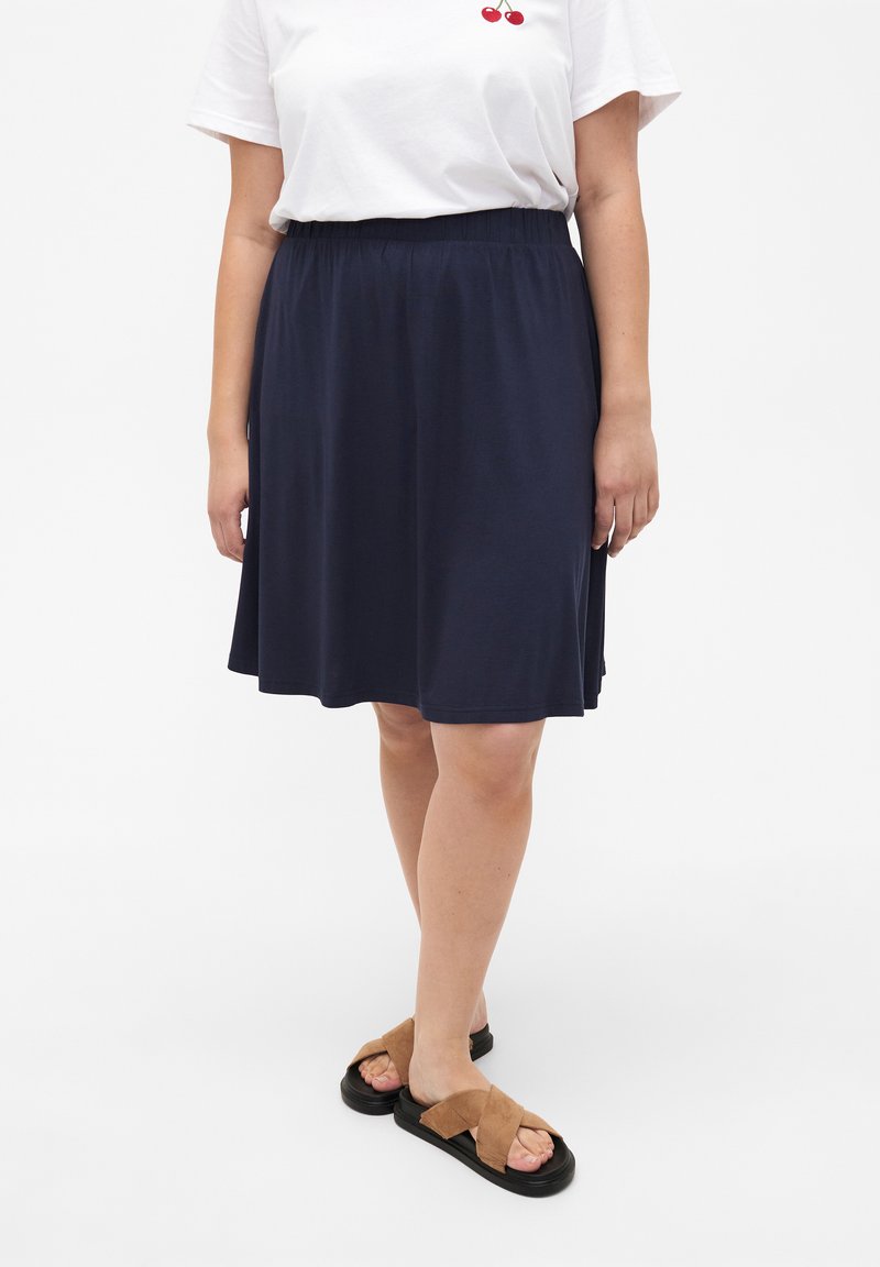 Jupe bleu marine à mi-mollet en tissu doux avec une taille élastique. Associée à un t-shirt blanc et des sandales croisées marron.