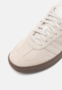 Beige suede sneaker with flat laces, rounded toe, and textured brown rubber sole featuring small spikes along the edge.