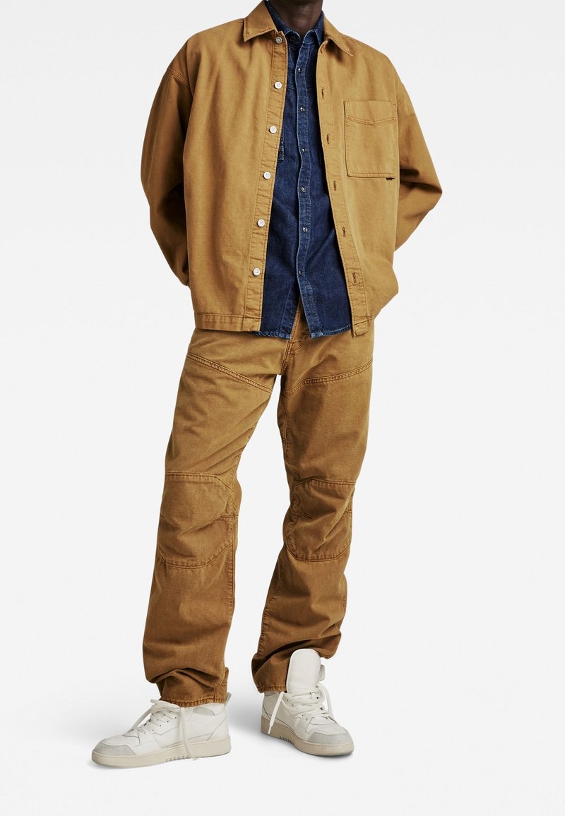 Brun canvasjacka över en mörkblå denimskjorta, i kombination med tan cargo-byxor och vita sneakers, med en avslappnad passform och synlig sömnad.