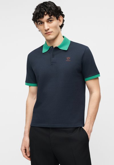 Νέος άνδρας με σγουρά μαλλιά, που φορεί ένα μπλε-σκούρο πόλο πουκάμισο με πράσινο γιακά και μανσέτες, στέκεται με το ένα χέρι στην τσέπη, μπροστά σε απλό φόντο.